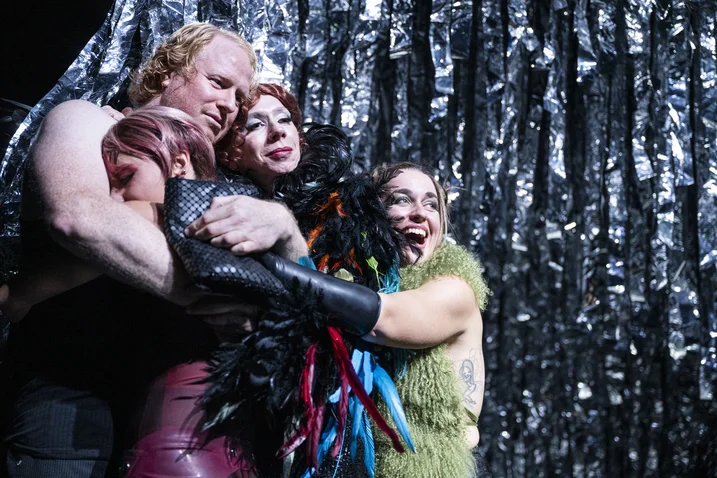 Three people in colorful costumes embrace in front of a shimmering silver backdrop. Expressive and emotional performance.