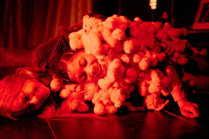 Eine Person ist mit vielen Kuscheltieren bedeckt, in warmem roten Licht getaucht. Die Szene wirkt gemütlich und verspielt.