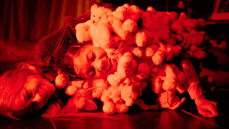 A person is covered with numerous stuffed animals, bathed in warm red light. The scene appears cozy and playful.