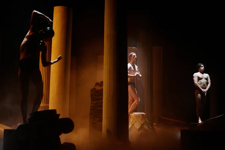 Eine Bühnenaufführung mit mystischer Beleuchtung zeigt Tänzer in antiker Kulisse zwischen hohen Säulen. Die Atmosphäre ist geheimnisvoll und ausgefallen.