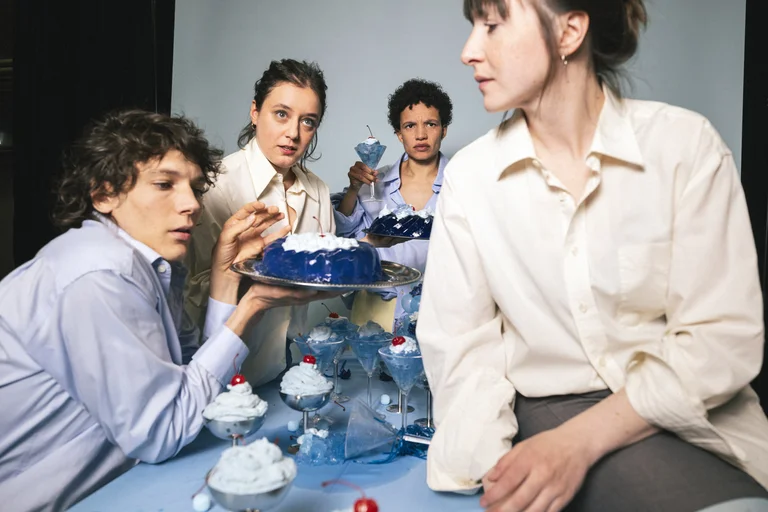 Four people are seated at a table filled with dessert dishes, wearing light clothing, looking in various directions.