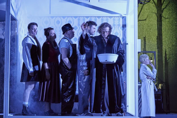 Actors on stage in a surreal, blue-lit scene. Some stand in a line while one person is in the background.