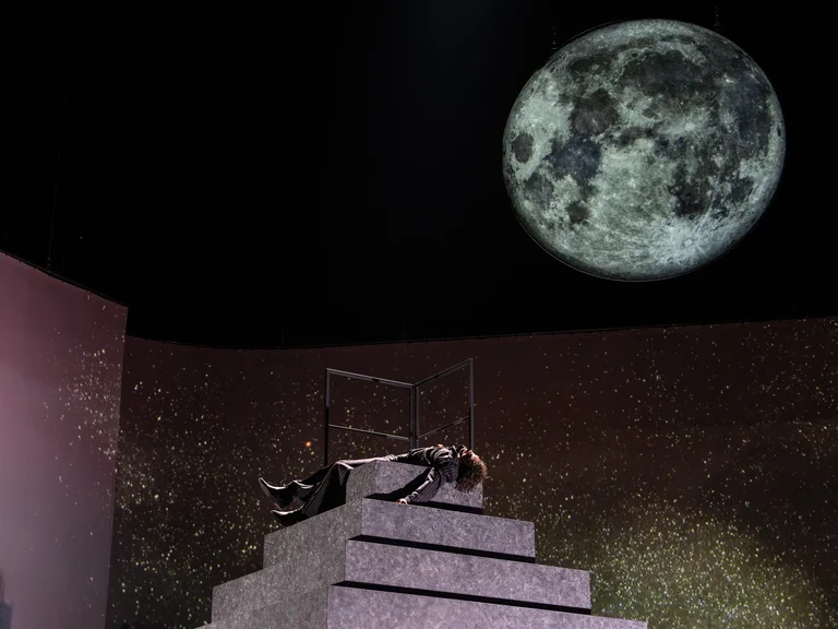 Ein Bühnenbild mit einer großen Nachbild des Mondes. Eine Person liegt auf einer Treppe, die Szene ist dramatisch beleuchtet.