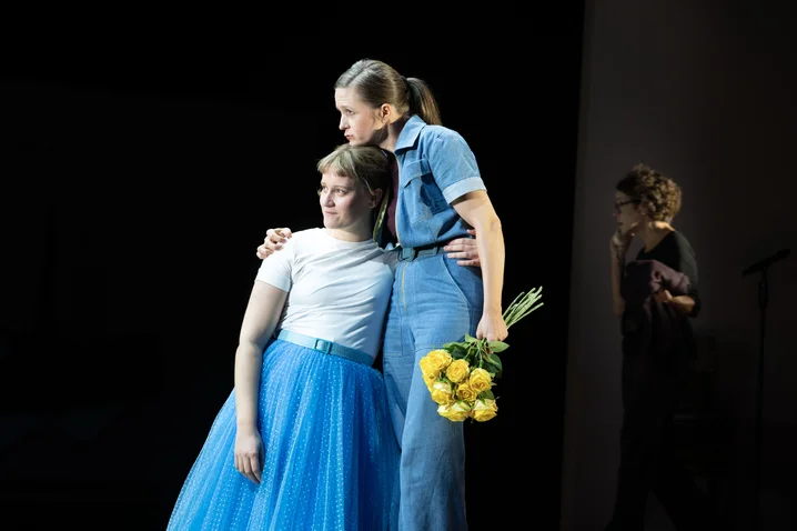 Two women on stage share a gentle embrace. One wears a blue skirt; the other holds yellow flowers in hand.