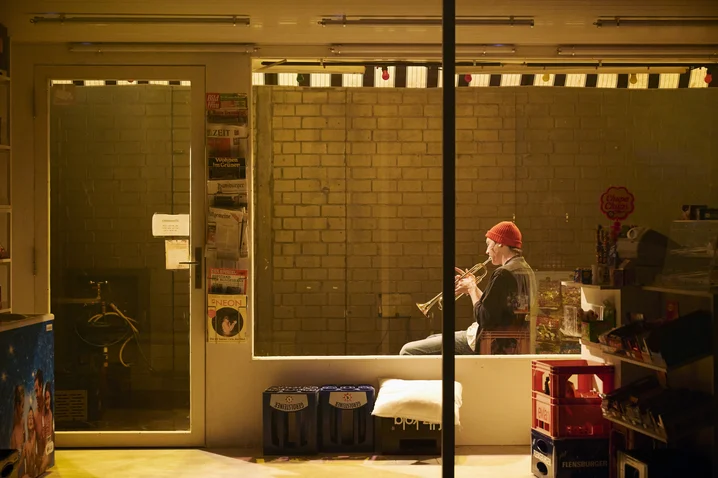 A musician plays the flute in a shop window. The scene is warmly lit, creating a peaceful evening atmosphere.