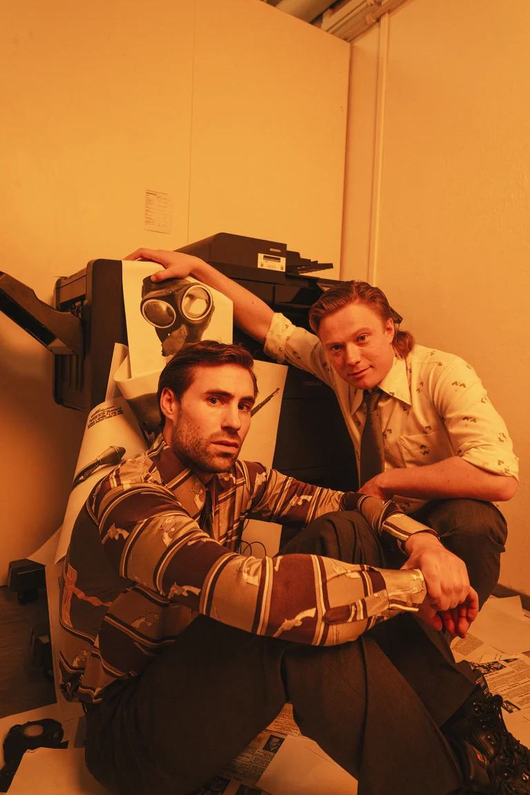 Two men pose in an orange-lit room beside a retro-futuristic machine. They are wearing patterned shirts.