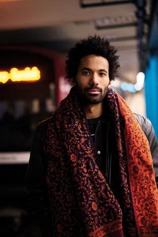 Eine Person mit Afrohaarschnitt trägt einen dekorativen, roten Schal in einer U-Bahn-Station. Im Hintergrund sind verschwommene Lichter sichtbar.