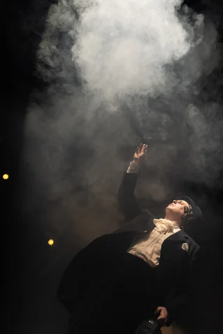 A person in an elegant black coat stands beneath a cloud of smoke, dramatically reaching an arm upwards.