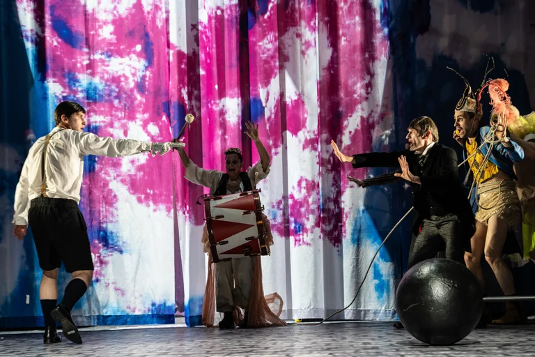 Eine theatralische Aufführung mit Darstellern auf der Bühne. Im Hintergrund sind bunte Vorhänge, vorne interagieren Personen in Kostümen miteinander.