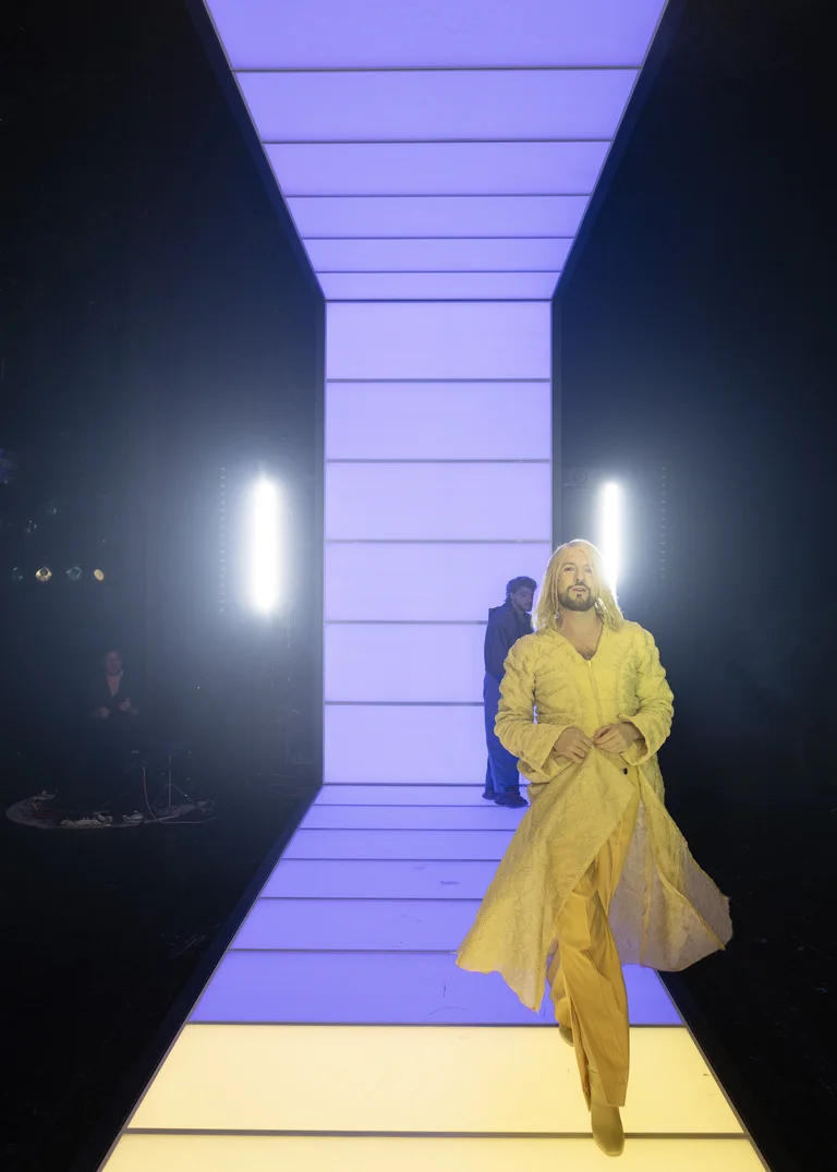 Model in a yellow outfit walks down a brightly lit runway. The background is illuminated with purple and white lights.